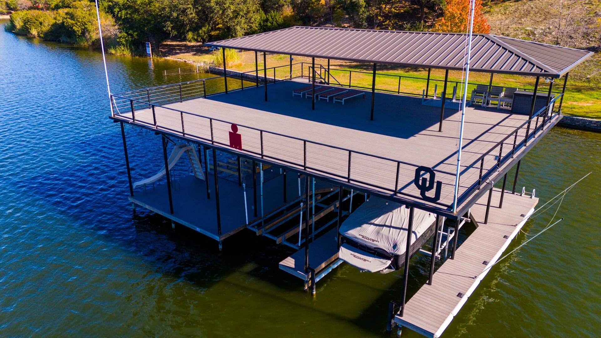 Aerial view of a modern covered boat dock on Lake Granbury featuring a spacious upper sundeck with composite decking and metal railings. The dock includes a covered boat slip with a stored vessel beneath a brown metal roof.