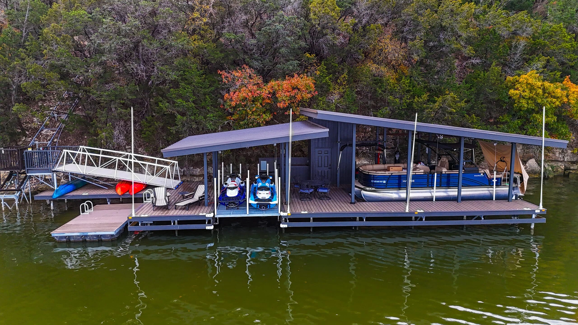 Waterfront boat dock on Lake Granbury featuring multiple vessel storage areas, including slips for two jet skis and a pontoon boat. The covered structure has a modern gray metal roof and composite decking, with a storage room in the center.