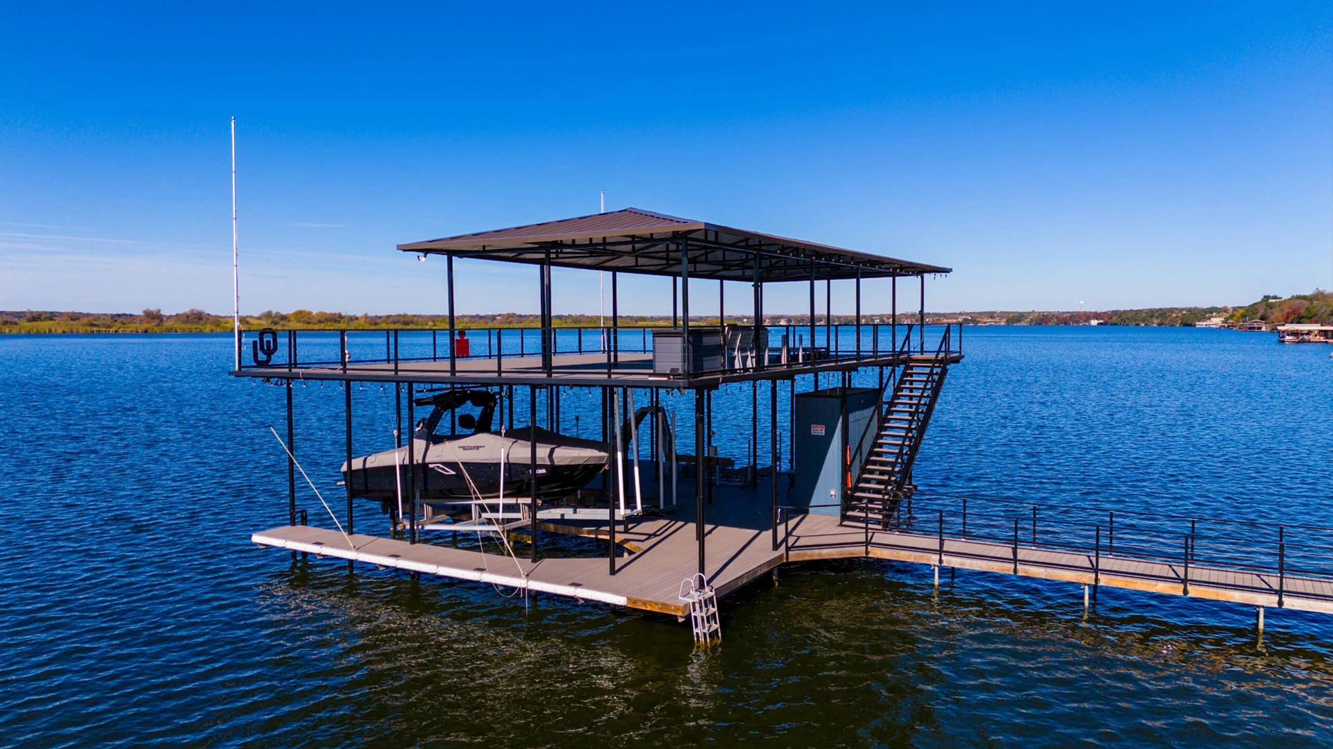 Two-story boat dock on Lake Granbury photographed on a clear day with vibrant blue skies and water. The dock features a covered upper deck with brown metal roofing and a boat lift below housing a white recreational vessel.