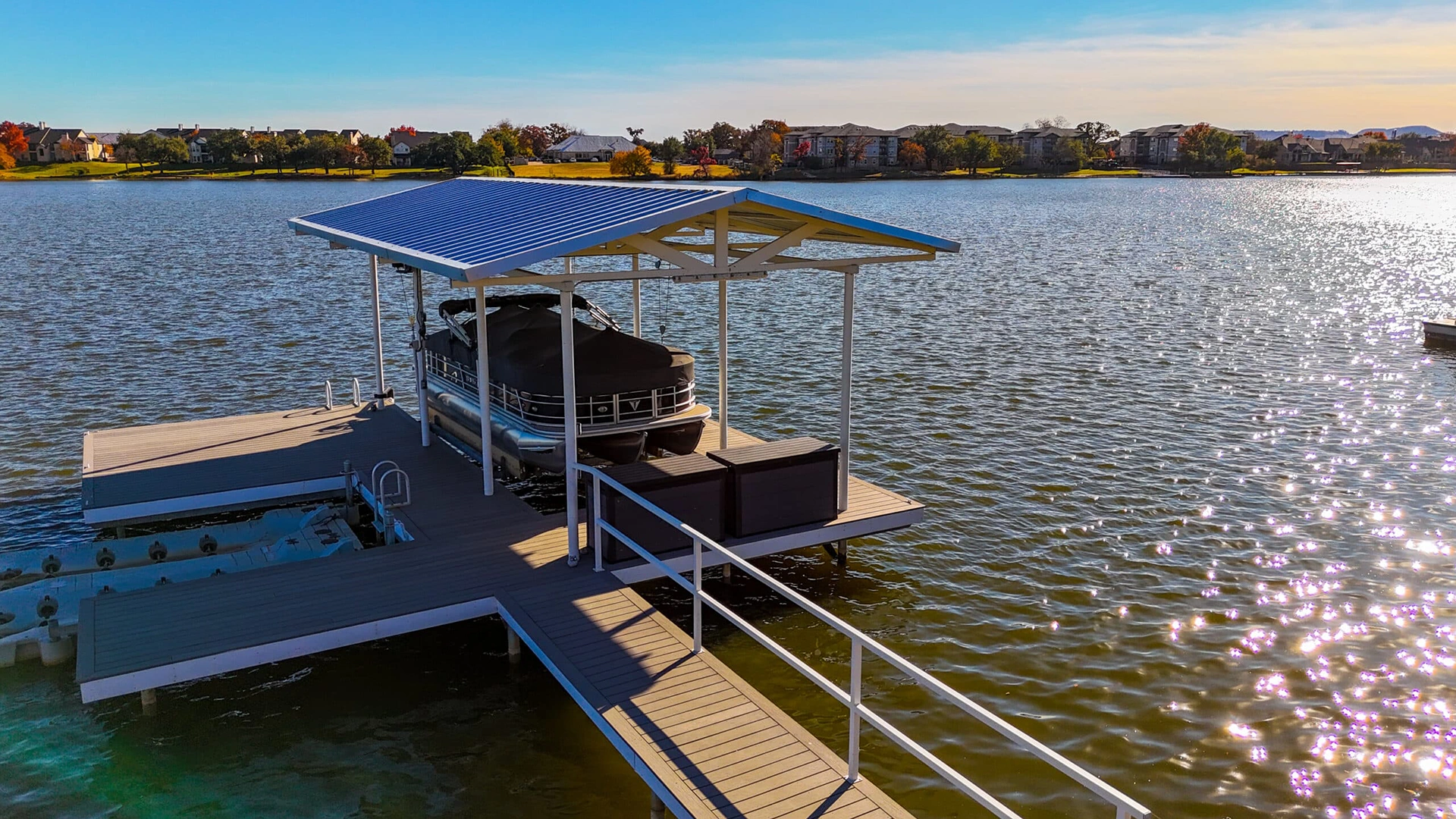 Lakefront dock on Lake Granbury with a covered pontoon boat slip, featuring a blue metal roof and white support columns. The composite decking creates an L-shaped platform with storage boxes, and white safety railings line the walkway.