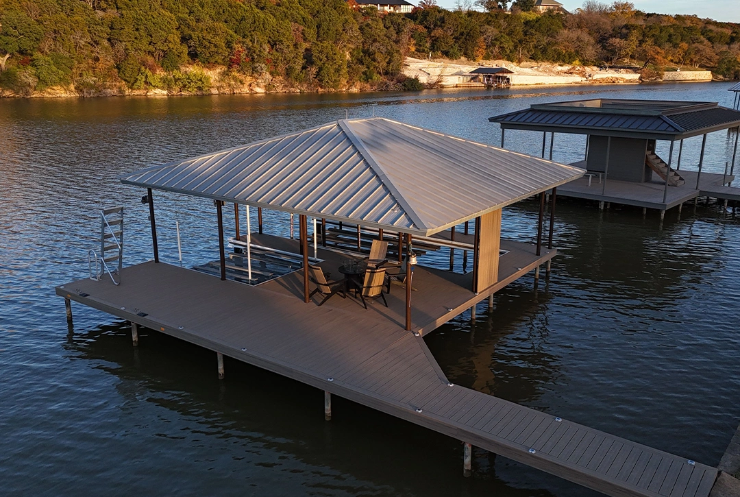 A custom boat dock extends into Lake Granbury with a modern design featuring a covered slip area with a metal hip roof. The dock has composite decking in a warm brown tone and includes a seating area with wooden Adirondack chairs.